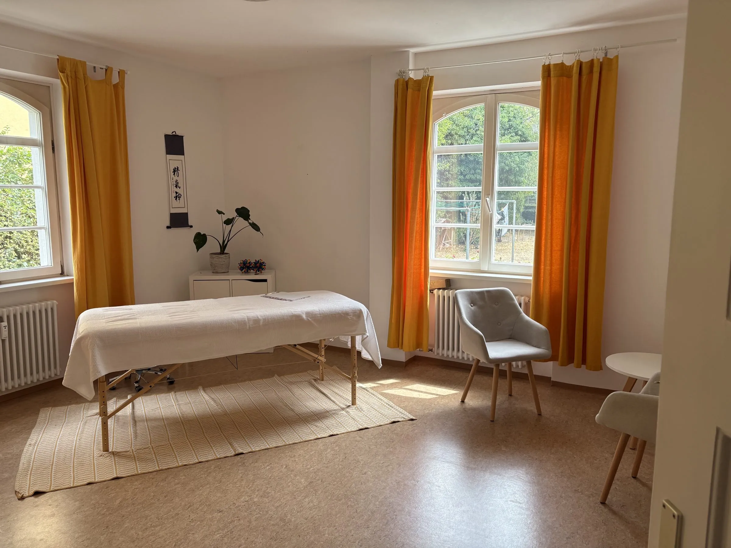 Die Behandlungsliege im Praxisraum, dahinter zwei Rundbogenfenster mit safrangelben Vorhängen