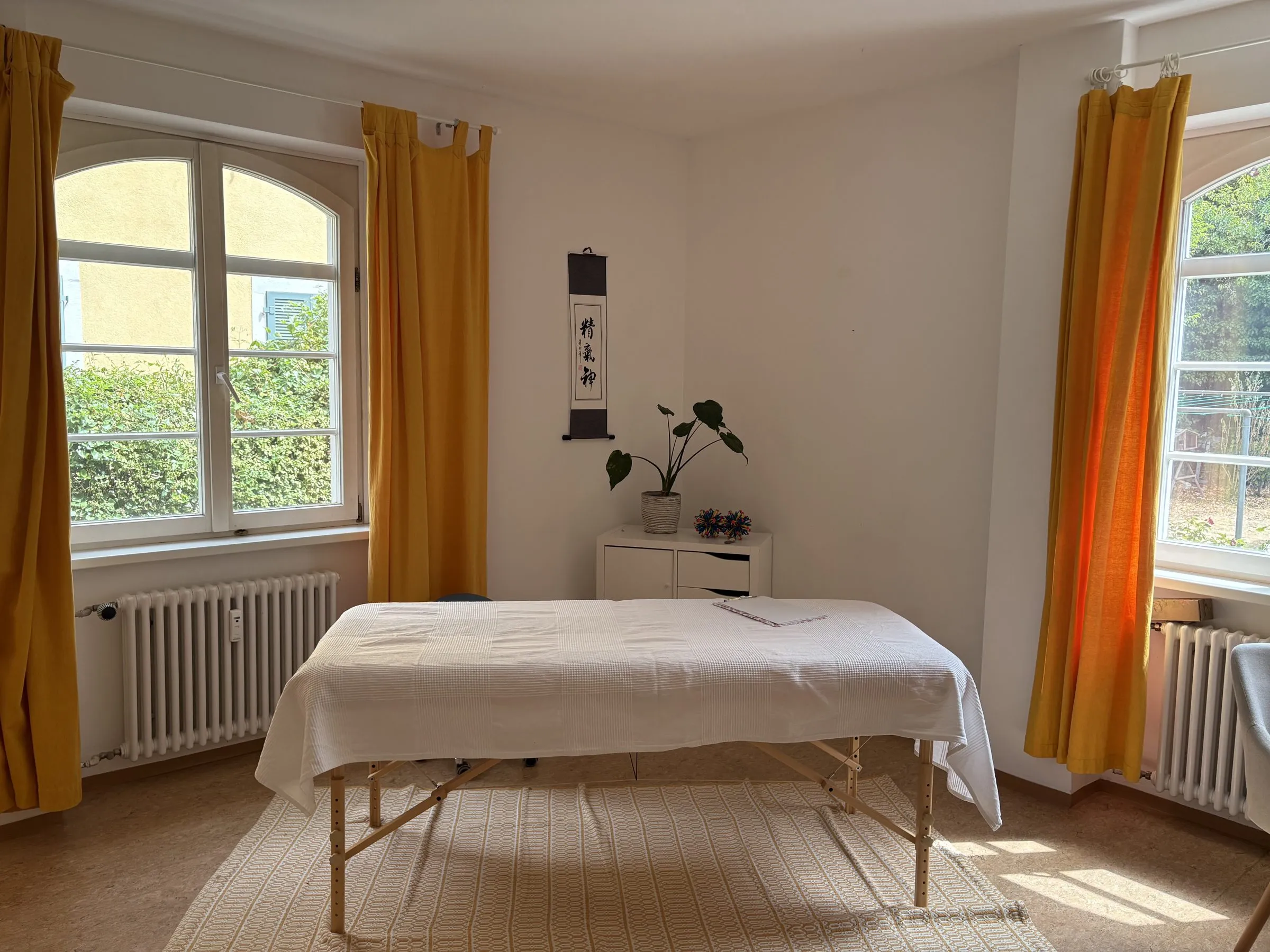Die Behandlungsliege mit weißer Decke zwischen zwei Fenstern